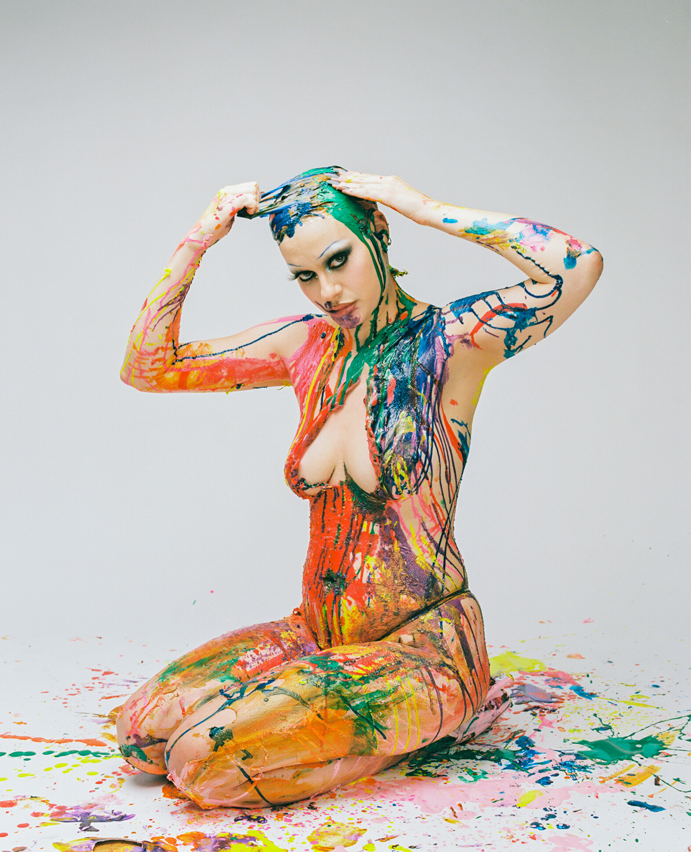 A Person with their skin covered in colorful paint splatters and streaks kneels on the ground serving the camera an intense gaze. The intentional composition of the paint splatter is apparent in how the paint frames their chest giving the appearance of cleavage.